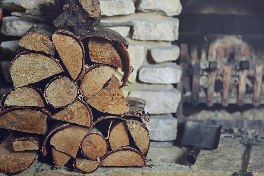 A stack of split firewood sits in front of a stone fireplace hearth next to a small fireplace shovel and poker.