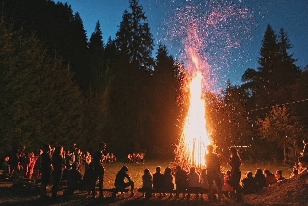 A roaring bonfire in a clearing surrounded by people and tall pine trees reaches high into the night sky, burning dried firewood.
