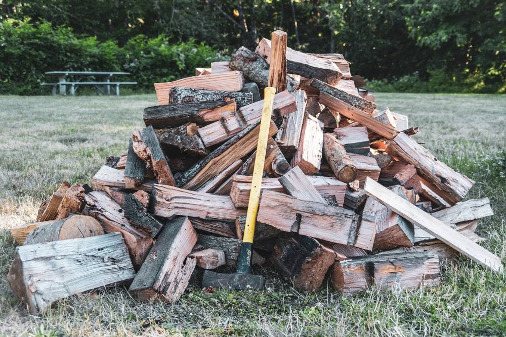 A three-foot tall pile of split firewood sits in a clearing with a yellow handled axe leaning against it and a green picnic table at the tree line in the background.